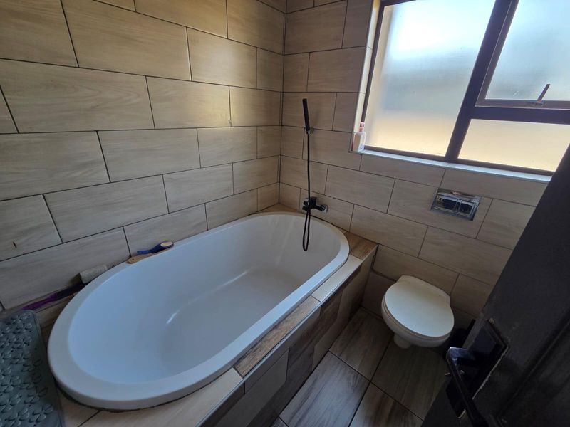 A modern bathroom featuring a built-in bathtub with wall-mounted shower fixture, a toilet, and neutral wood-look wall and floor tiling.