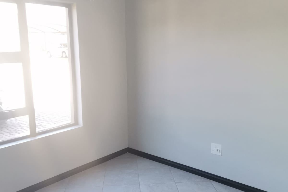A bright, empty bedroom featuring neutral grey walls, tiled flooring, and a large aluminium window providing natural light.