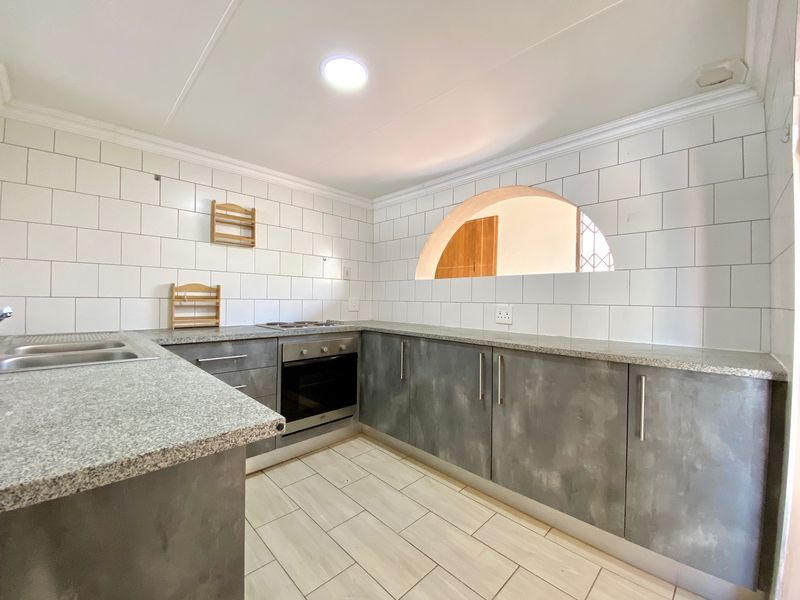 A clean, modern kitchen featuring grey cabinetry, granite countertops, subway tile backsplash, and a unique arched pass-through window to the adjacent living area.