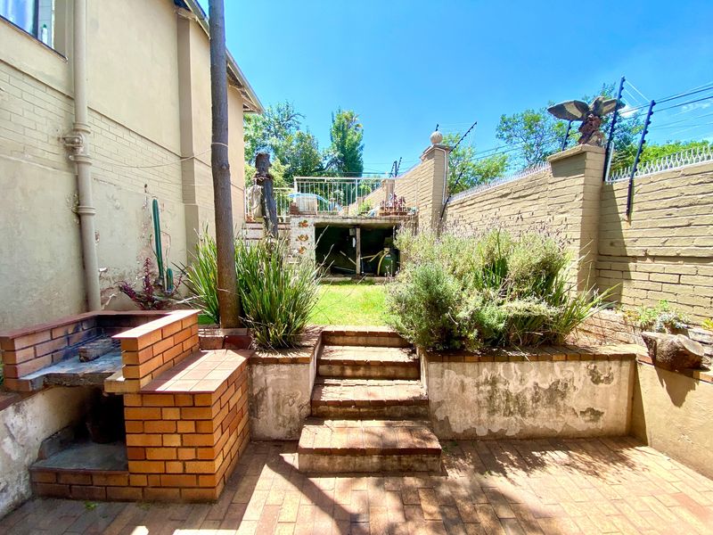 Rear garden area featuring a built-in brick braai, paved patio, tiered retaining walls, and secure perimeter boundary with electric fencing.