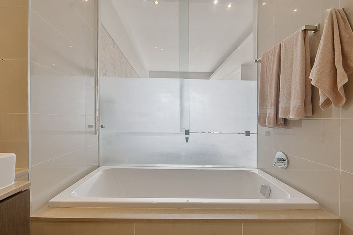 A modern bathroom featuring a built-in bathtub with a frosted glass screen, neutral wall tiling, and recessed ceiling lighting.