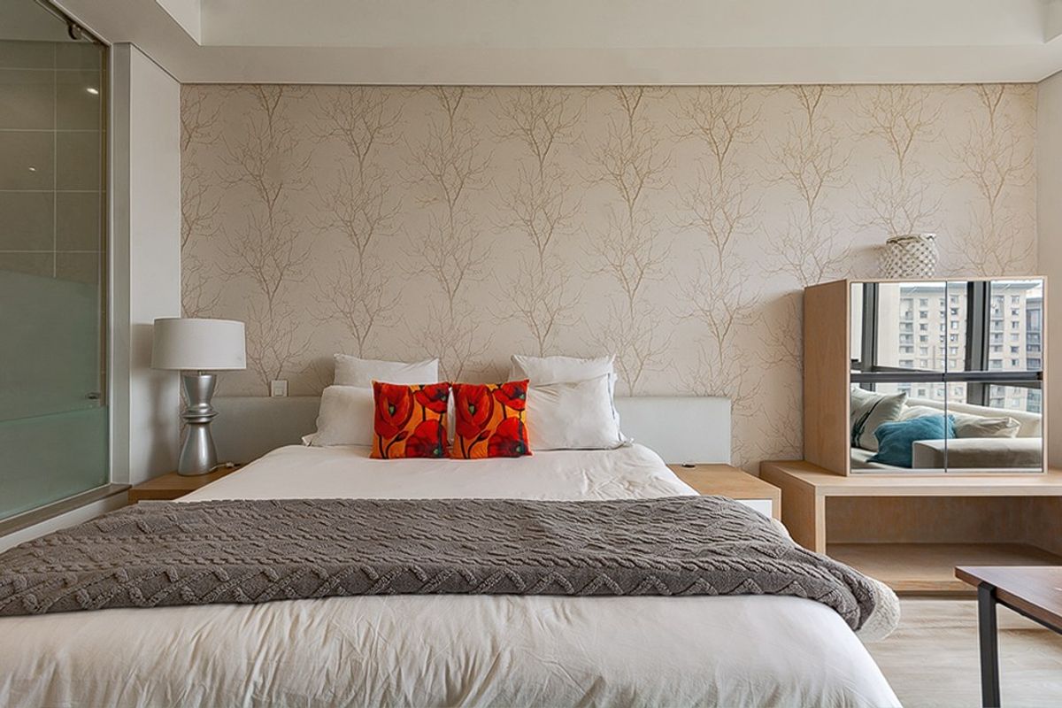 A well-maintained bedroom featuring a built-in headboard with integrated bedside tables, decorative wallpaper, and an adjacent glass-enclosed ensuite bathroom.