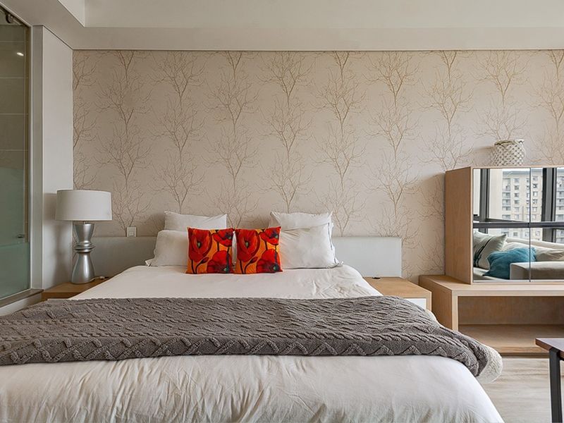 A well-maintained bedroom featuring a built-in headboard with integrated bedside tables, decorative wallpaper, and an adjacent glass-enclosed ensuite bathroom.