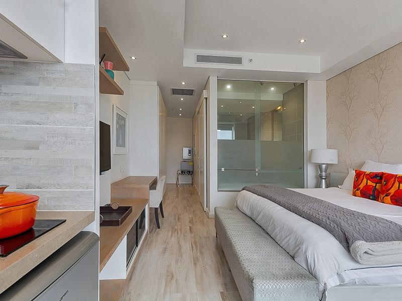 Modern open-plan studio apartment featuring a compact kitchen area, built-in desk, and a glass-enclosed bathroom, finished with light wood-look flooring and recessed lighting.