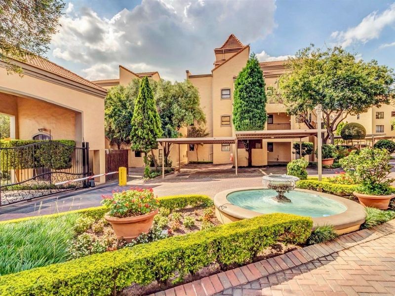 A well-maintained apartment complex entrance featuring a paved driveway, covered parking, landscaped gardens, and a decorative water fountain.