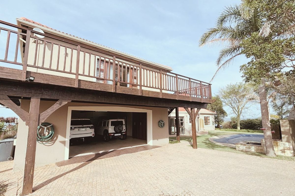 Rear exterior view featuring a raised timber deck, double garage, paved driveway, and a swimming pool with an outdoor fireplace area.