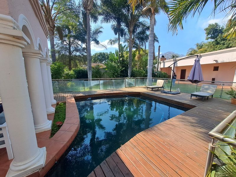 Outdoor pool area featuring timber decking, glass balustrades, and classical architectural columns, set within a landscaped garden environment.