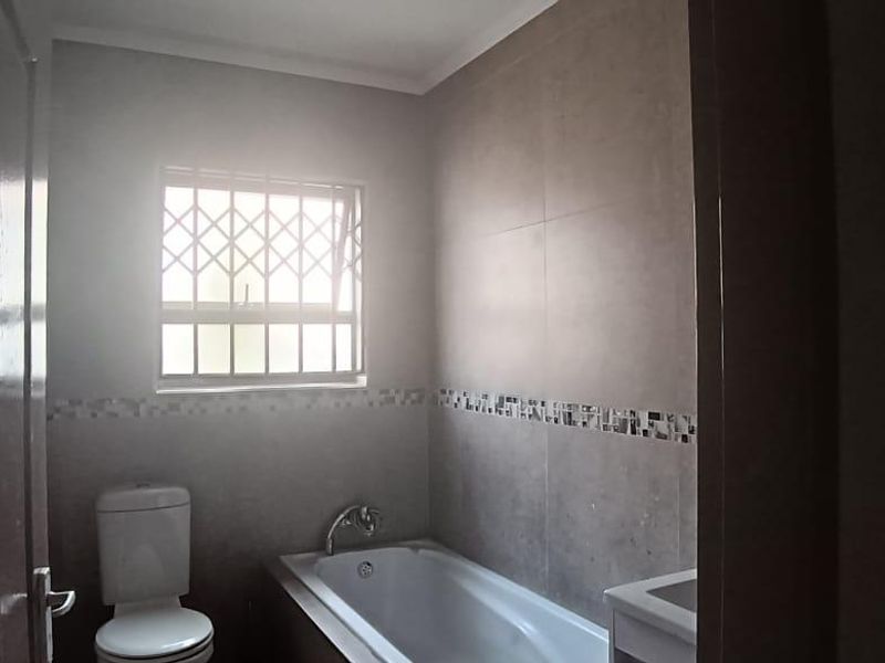 A clean, functional bathroom featuring a bathtub, toilet, and vanity, with neutral wall tiling and a window fitted with security bars.