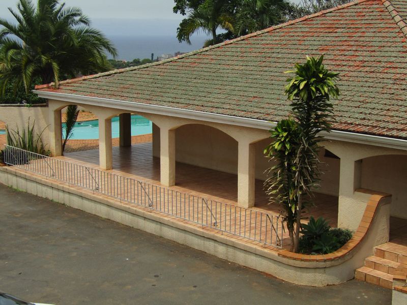 A covered patio area featuring arched pillars, tiled flooring, and a metal railing, overlooking a swimming pool with distant sea views.
