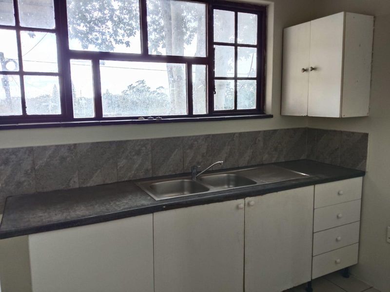 A functional kitchen area featuring a double bowl stainless steel sink, laminate countertops, tiled backsplash, and ample built-in cabinetry under a large window.