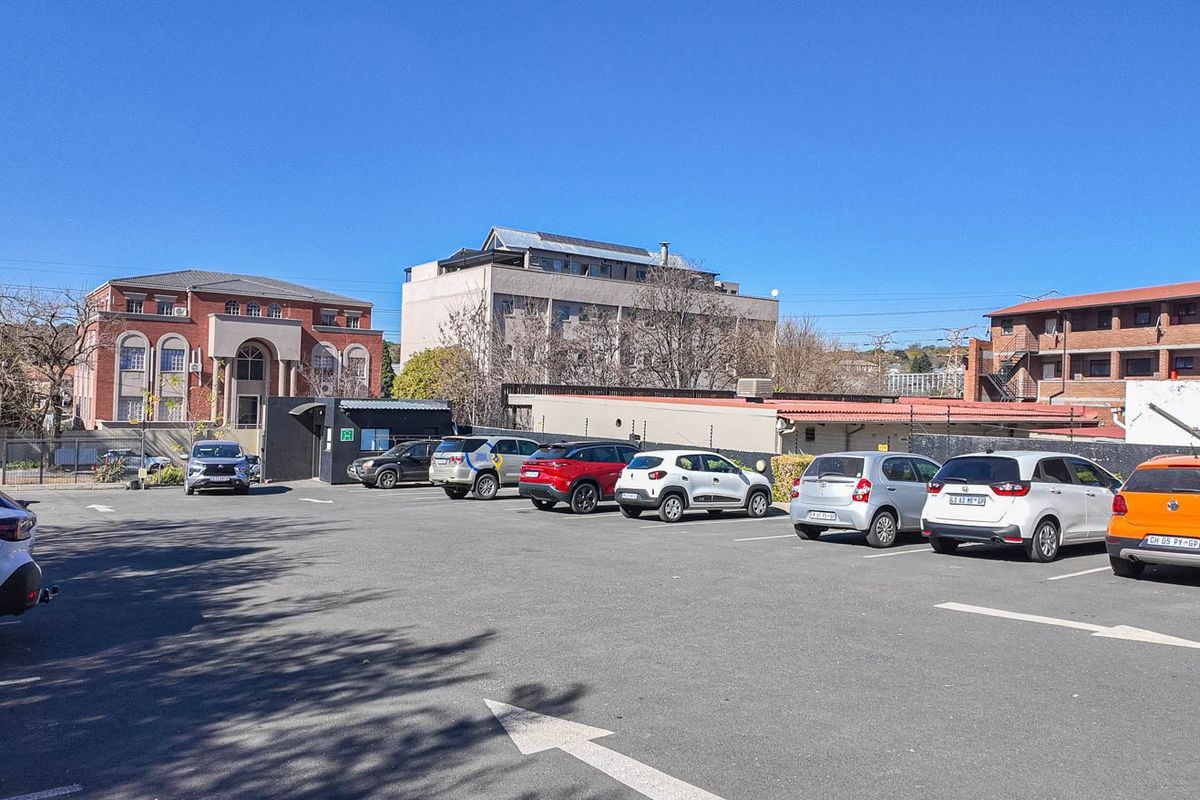 A paved commercial parking area featuring marked bays, perimeter security fencing, and access to surrounding multi-story office and residential buildings.