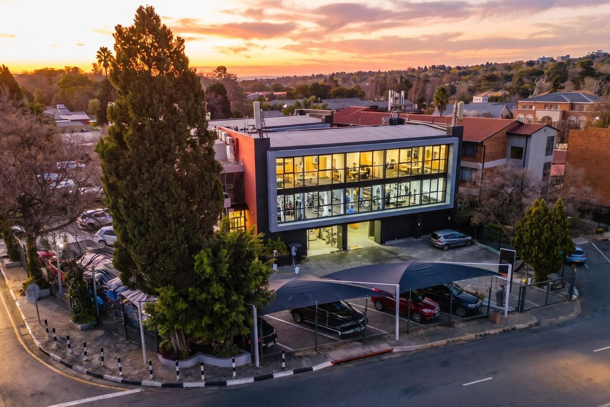 Aerial view of a modern multi-story commercial building featuring a large glass facade, covered parking facilities, and surrounding landscaped grounds at sunset.