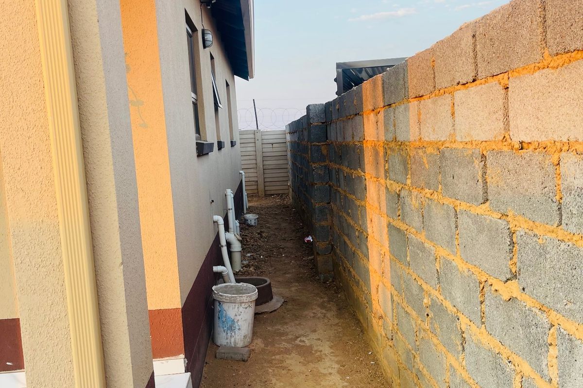 Side passage of the property featuring a concrete block boundary wall, external plumbing infrastructure, and an unpaved ground surface.