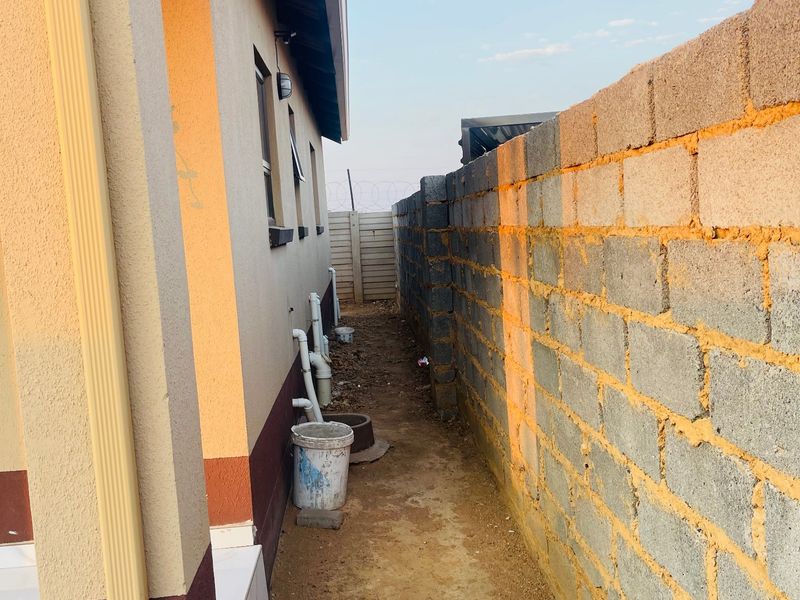 Side passage of the property featuring a concrete block boundary wall, external plumbing infrastructure, and an unpaved ground surface.