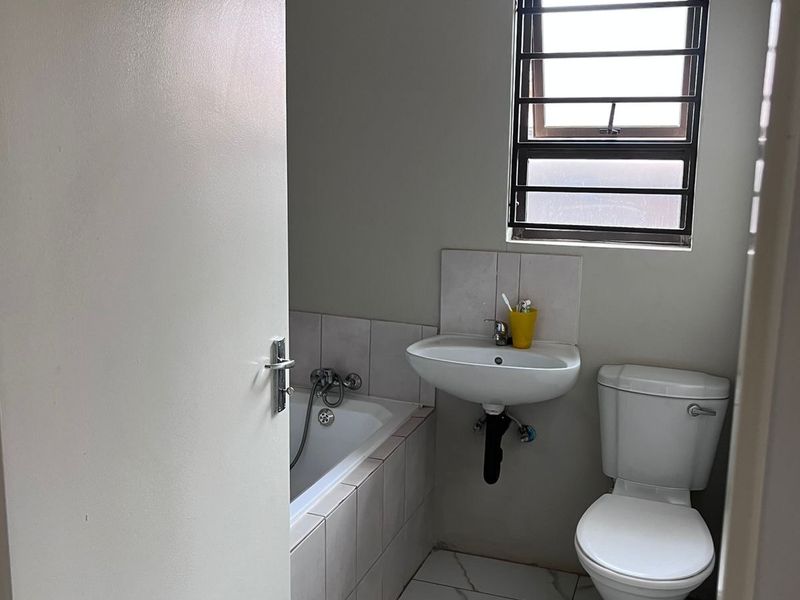 A functional bathroom featuring a bathtub, wall-mounted basin, and toilet, finished with modern marble-look floor tiles and a window with security bars.
