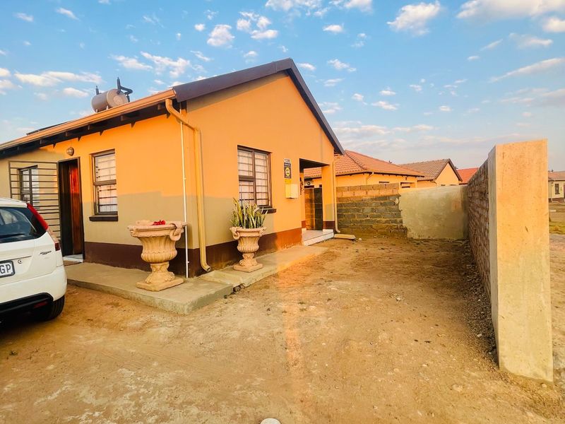 Front view of a single-story residential house featuring a solar geyser, security bars on windows, and an unpaved yard area with a boundary wall.