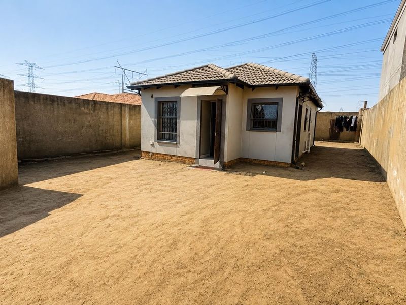 A rear view of a detached house featuring a tiled roof, security bars on windows, and a large, unlandscaped yard enclosed by perimeter walls.