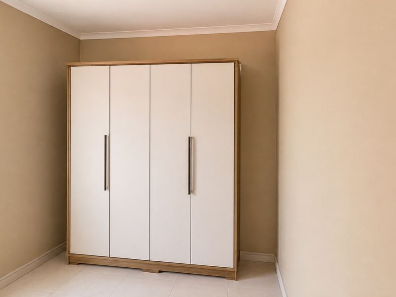 A clean, neutral bedroom featuring a modern built-in wardrobe with light wood trim and white doors, set against beige walls and tiled flooring.