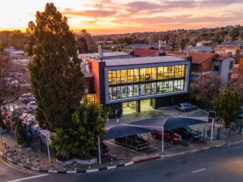 Aerial view of a modern multi-story commercial building featuring a large glass facade, covered parking facilities, and surrounding landscaped grounds at sunset.