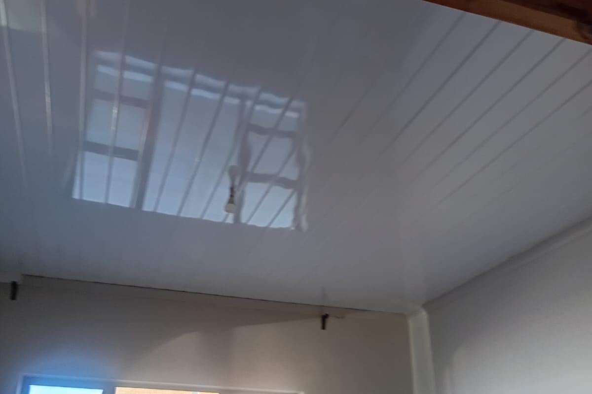 An empty bedroom featuring a glossy PVC panel ceiling with an exposed timber beam and aluminium window frames, currently requiring minor finishing work.