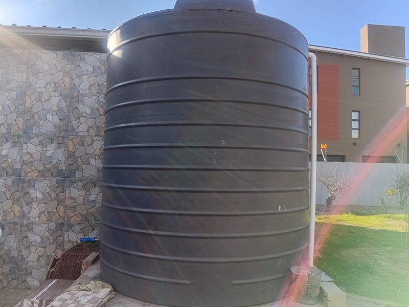 A large 5000-liter water storage tank installed on a paved area against a stone-clad exterior wall.