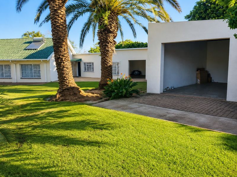 Rear view of the property featuring a green metal roof with solar geyser, open garage, paved driveway, and well-maintained lawn with mature palm trees.