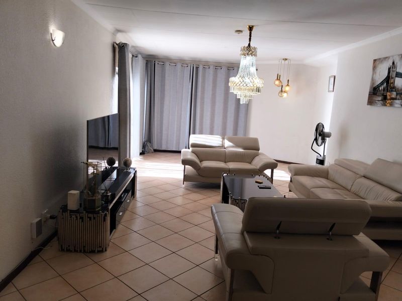Spacious living room featuring neutral tiled flooring, a decorative crystal chandelier, and large windows with curtain tracks, offering a bright and comfortable living space.