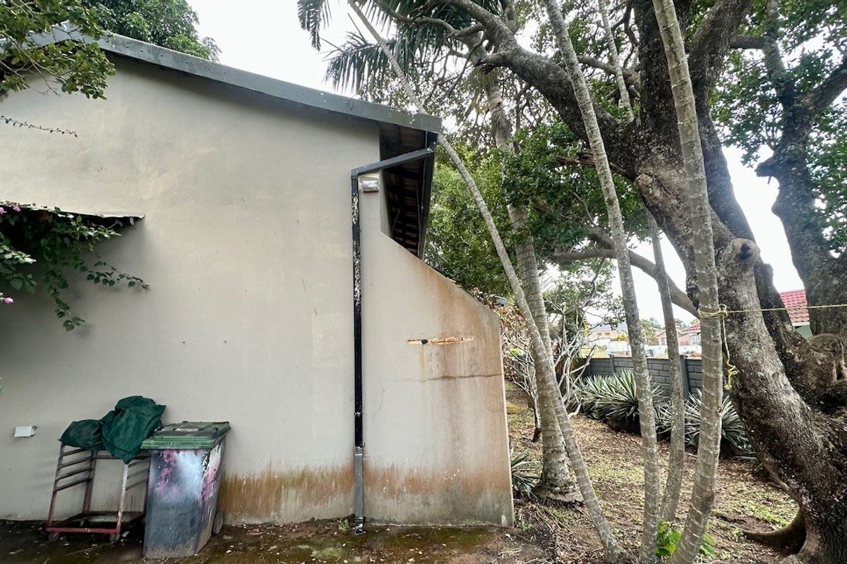 Side view of the property exterior showing masonry wall, rainwater downpipe, and surrounding garden area with mature trees.