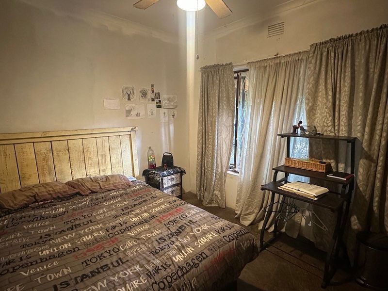 A bedroom featuring a ceiling fan and natural light from a window with curtains, requiring cosmetic updates to the wall finishes.