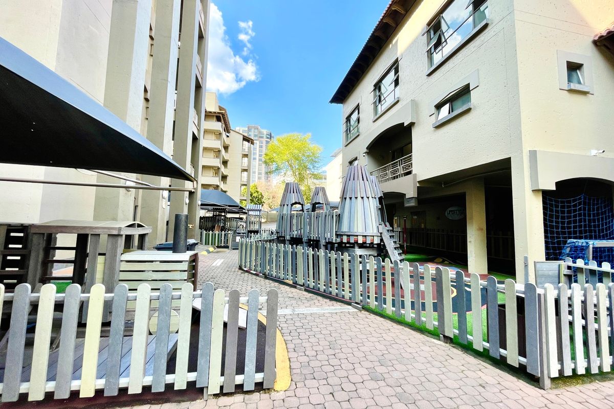 A communal outdoor area between apartment buildings featuring a paved walkway and a fenced playground facility.