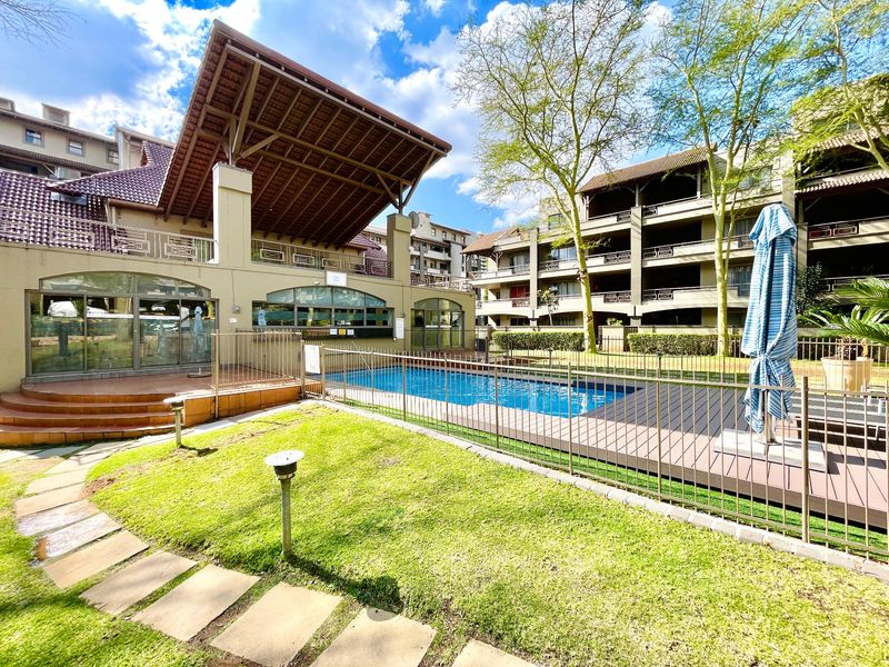 Communal swimming pool area featuring a paved deck, metal safety fencing, and landscaped garden surrounds within an apartment complex.