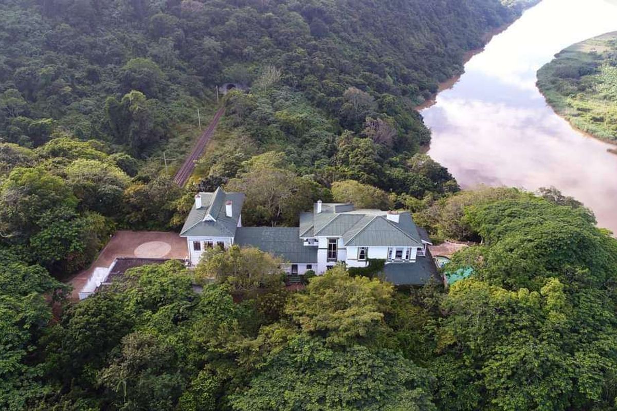 Aerial view of a large residential property nestled in lush greenery, featuring a multi-pitched roof, private driveway, and swimming pool overlooking a river.