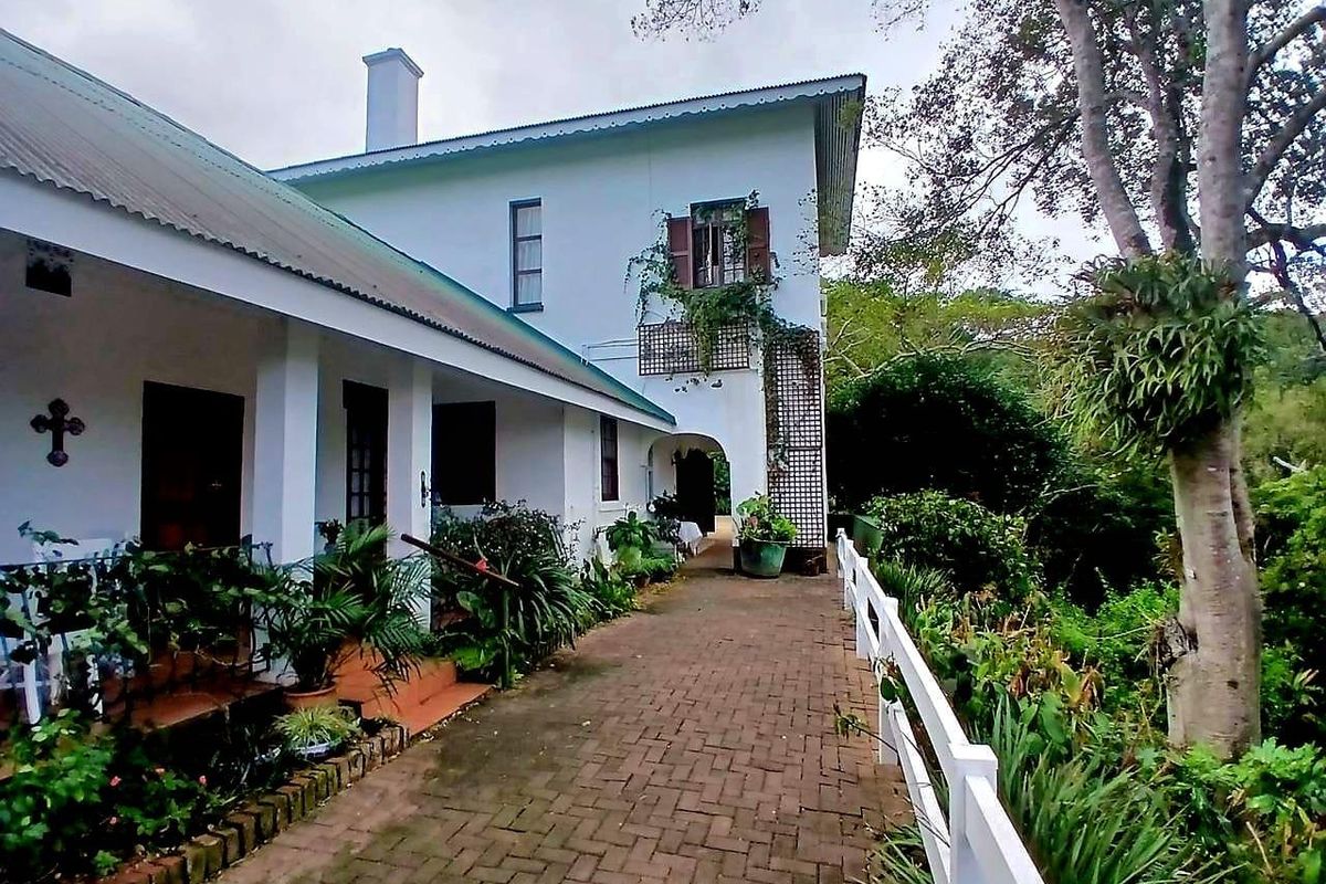 A charming rear exterior view featuring a covered patio, brick paved walkway, and a white picket fence set against a lush garden backdrop.