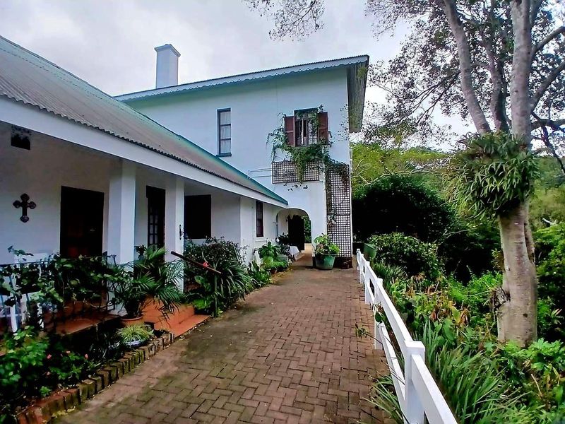 A charming rear exterior view featuring a covered patio, brick paved walkway, and a white picket fence set against a lush garden backdrop.