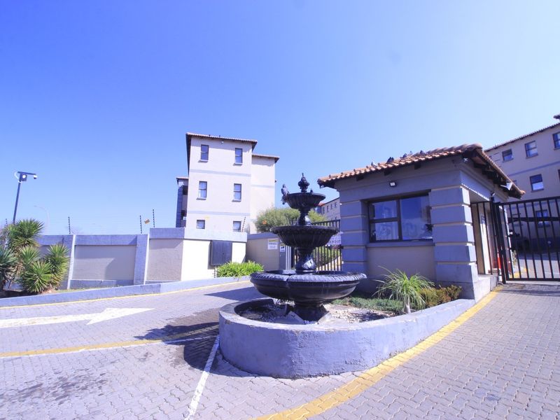 The apartment complex entrance features a secure gated access point, a guard house, and a decorative fountain set within a paved driveway area.