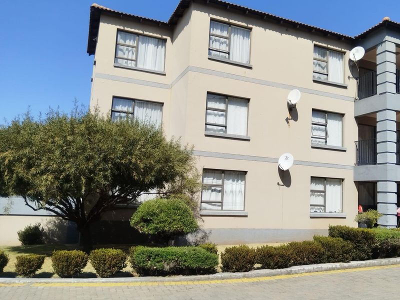 A three-story apartment building exterior featuring a beige facade, aluminium window frames, and a paved driveway with established garden landscaping.