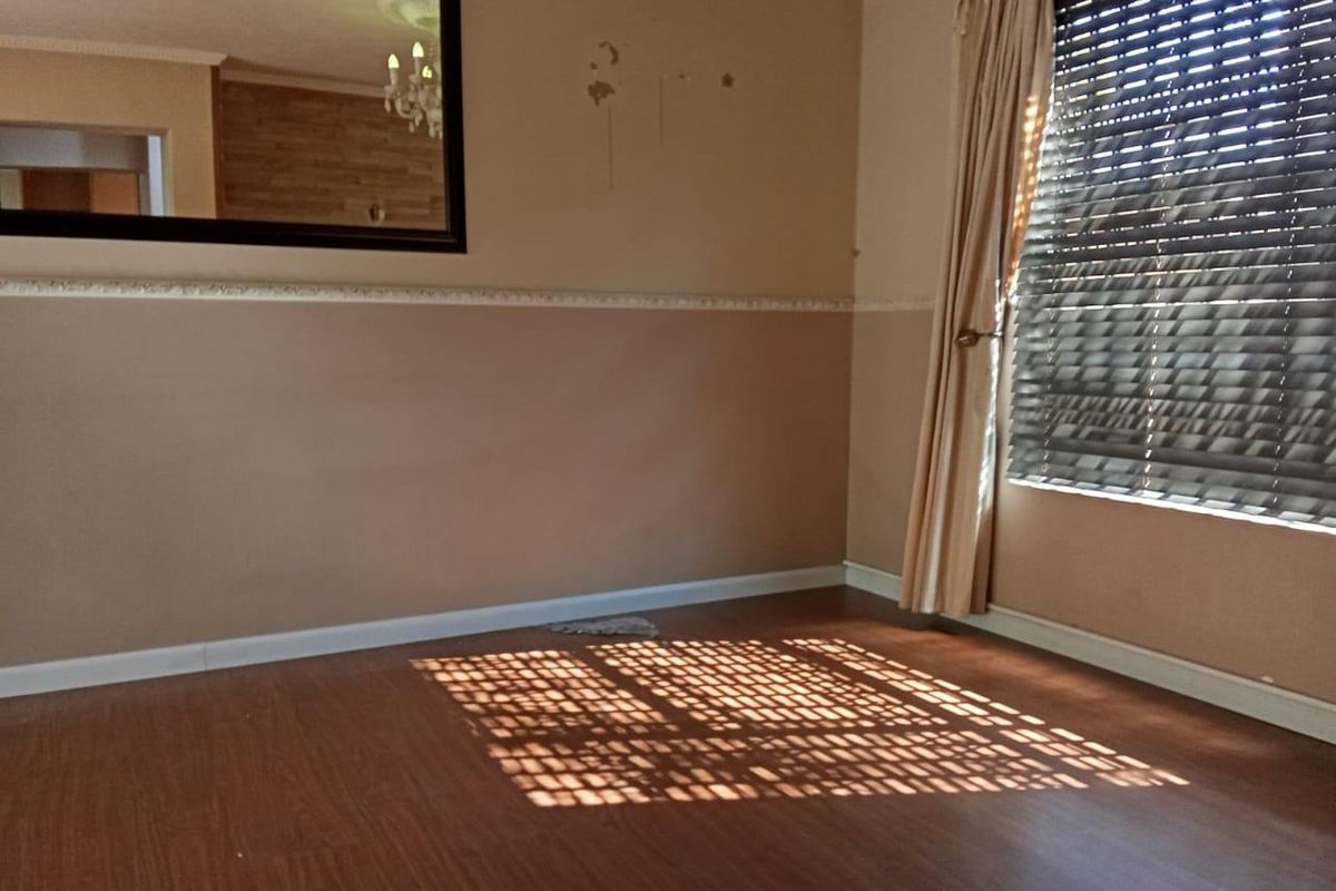 An empty living room featuring laminate flooring, a dado rail, and venetian blinds, showing signs of wear on the wall surfaces.