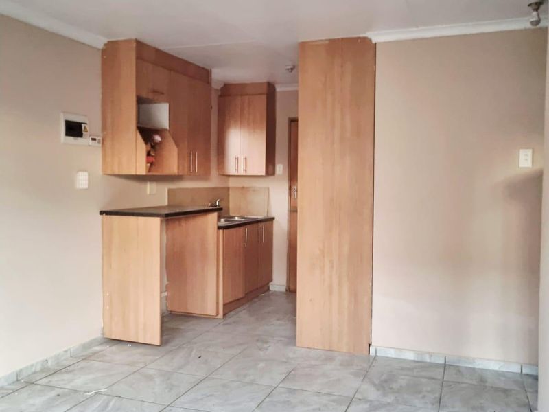 A compact, empty kitchen featuring light wood-finish cabinetry, laminate countertops, a stainless steel sink, and neutral tiled flooring.
