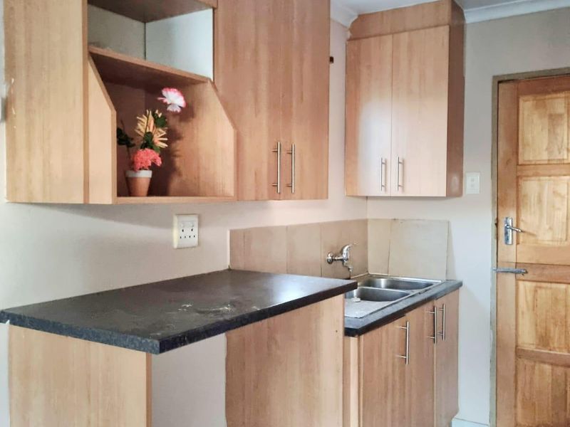 A compact kitchen featuring light wood-finish cabinetry, dark laminate countertops, a stainless steel sink, and tiled flooring with a wooden door.
