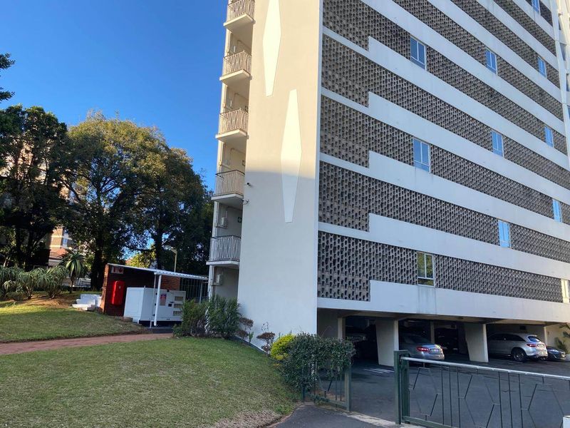 Exterior view of a multi-story apartment building featuring a distinctive breeze block facade, private balconies, and ground-level covered parking.