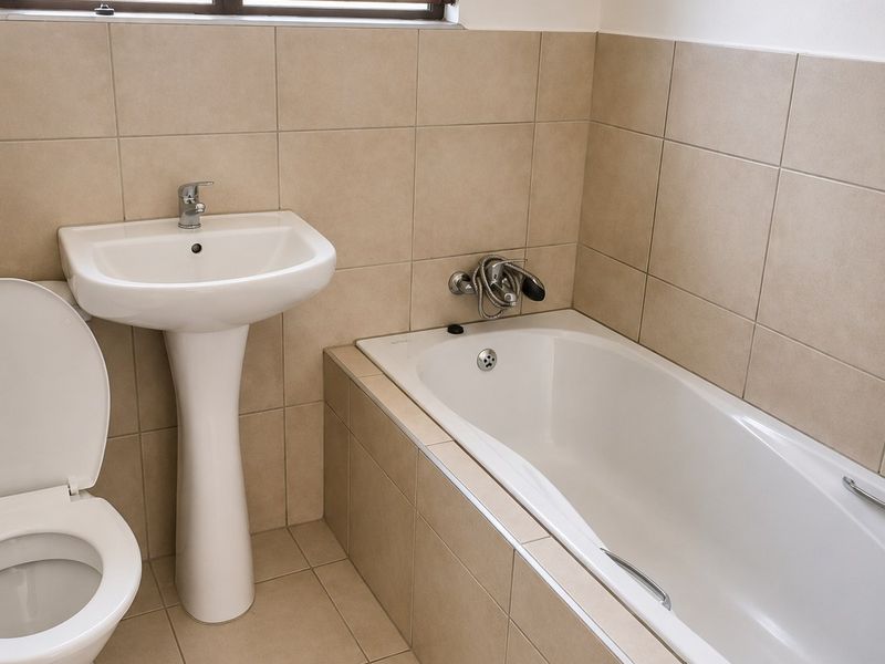 A clean, functional bathroom featuring a pedestal basin, built-in bathtub, and neutral wall and floor tiling with a window fitted with security bars.