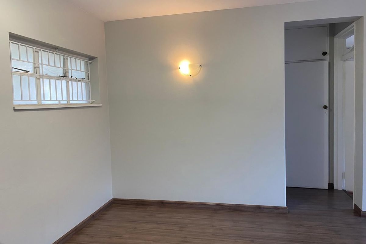 A clean, empty bedroom featuring laminate flooring, a wall-mounted light fixture, and a window with security bars.