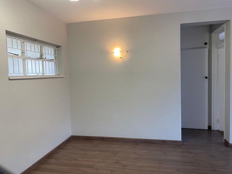 A clean, empty bedroom featuring laminate flooring, a wall-mounted light fixture, and a window with security bars.