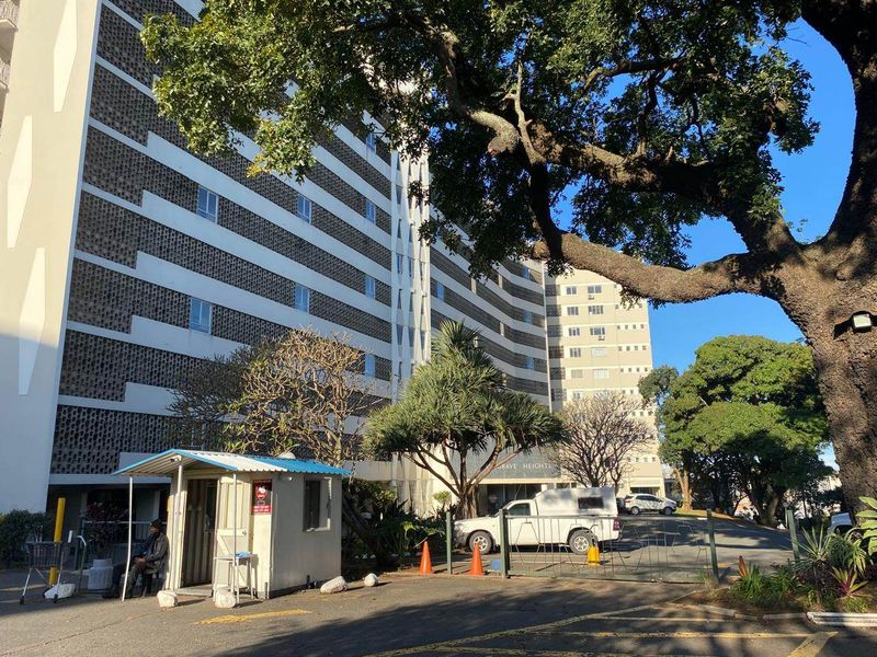 Exterior view of the apartment building entrance featuring a security guard booth, gated access, and mature trees.