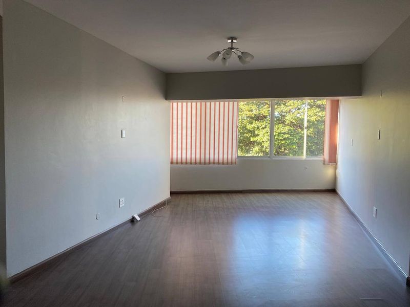 A spacious, empty living room featuring laminate flooring, neutral walls, and large windows with vertical blinds, offering a blank canvas for interior design.