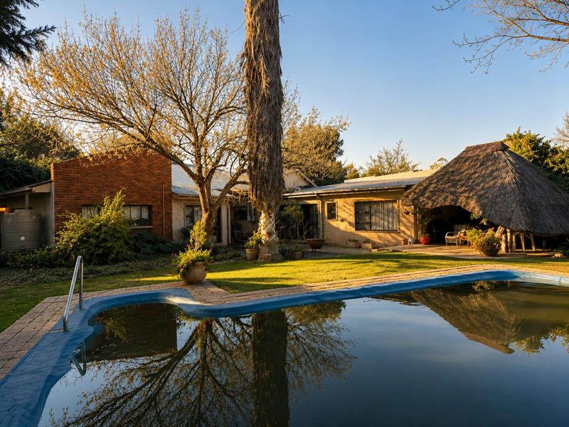 Rear garden view featuring a swimming pool with a pool cover, a thatched lapa entertainment area, and established trees.