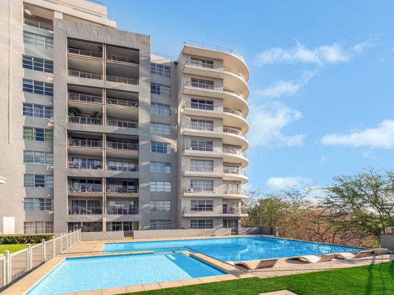 A well-maintained outdoor swimming pool area featuring a multi-level apartment building facade with private balconies in the background.