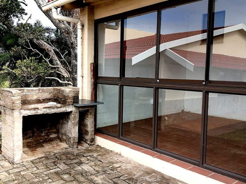 Covered patio area featuring a built-in brick braai, tiled flooring, and large aluminium-framed windows overlooking the garden.