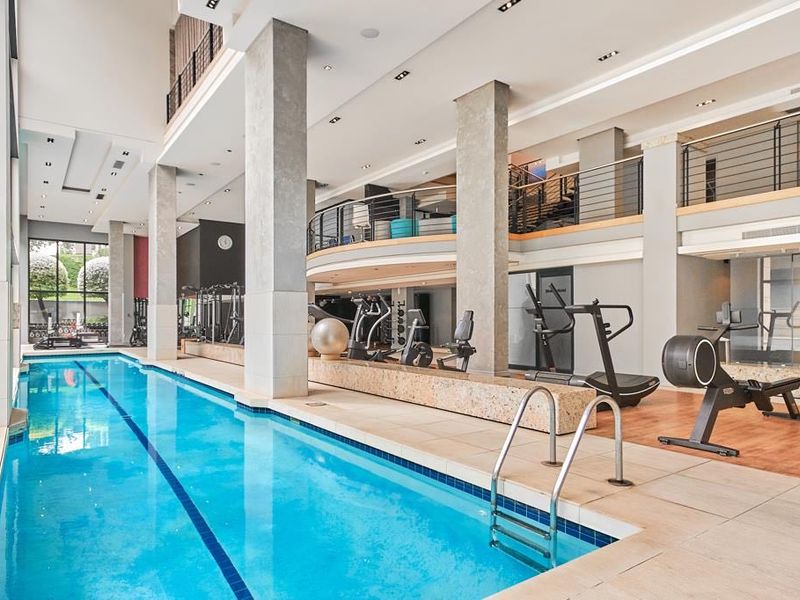 A modern indoor swimming pool area featuring high ceilings, concrete columns, and an overlooking mezzanine level with integrated gym facilities.