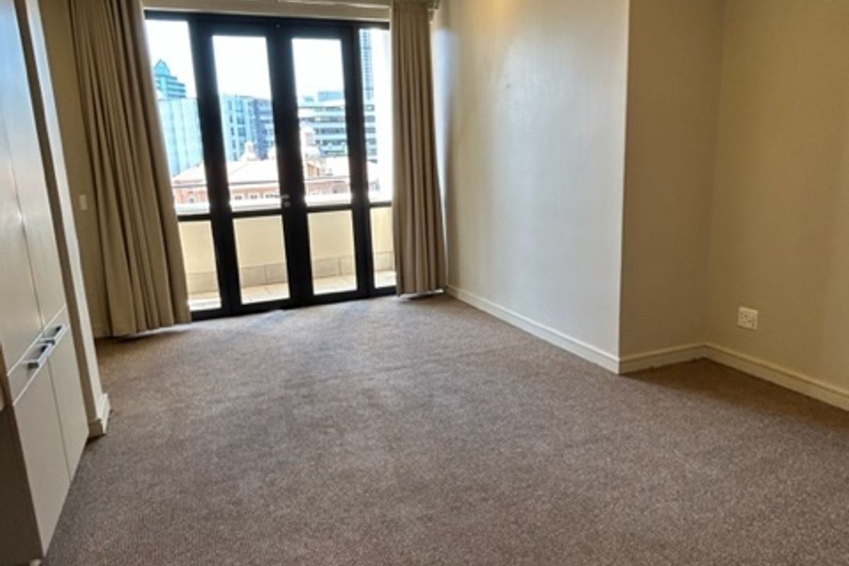 A spacious, empty bedroom featuring neutral carpeting, built-in cupboards, recessed lighting, and double glass doors leading to a balcony with city views.